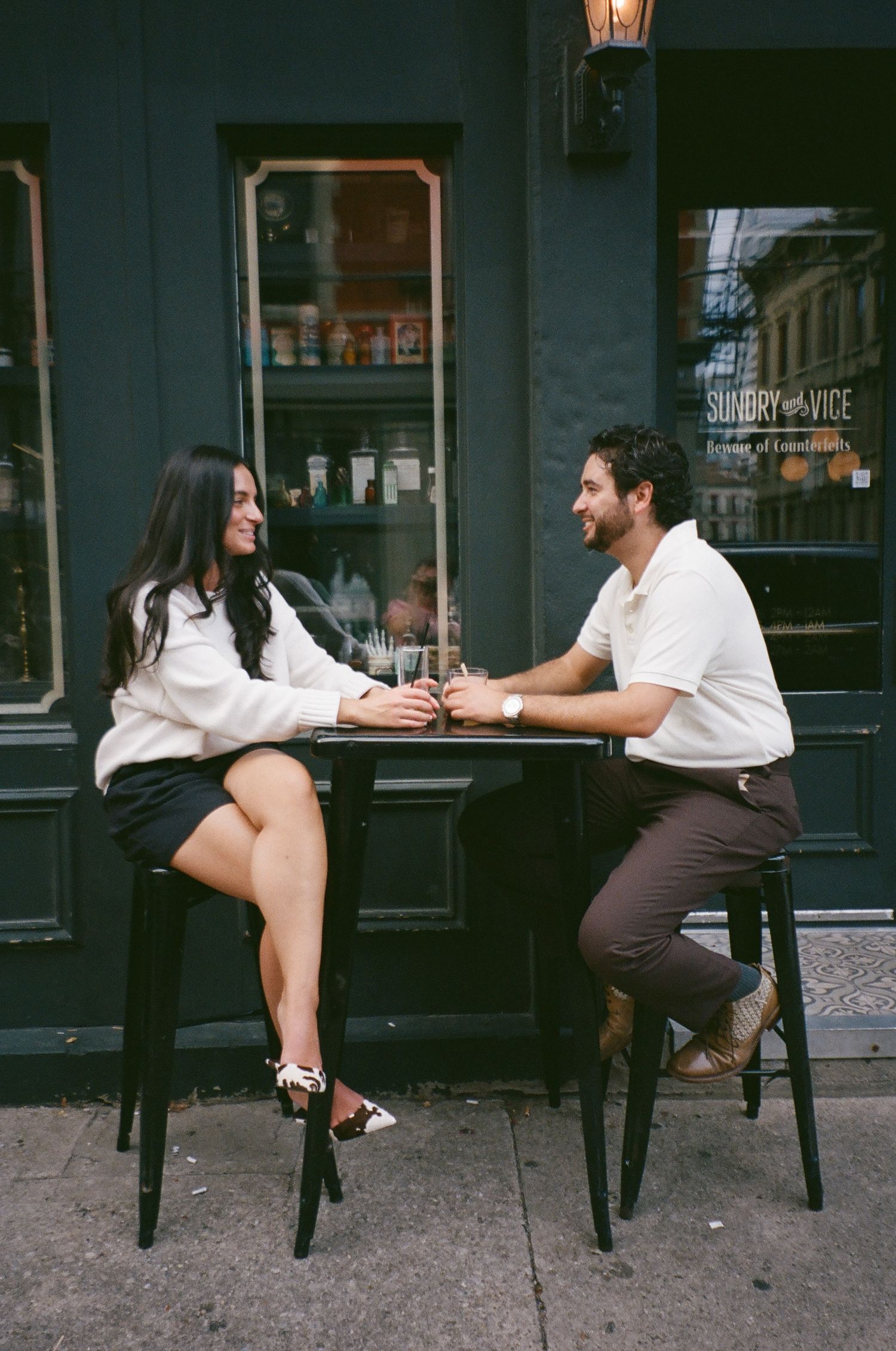 A couple dressed in summer white outfits stands outside a brick residence with lush greenery.
