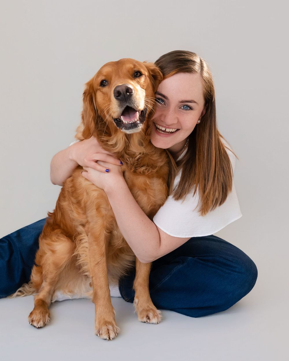 Young woman in white top and jeans smiling while hugging a happy golden retriever dog in a studio.