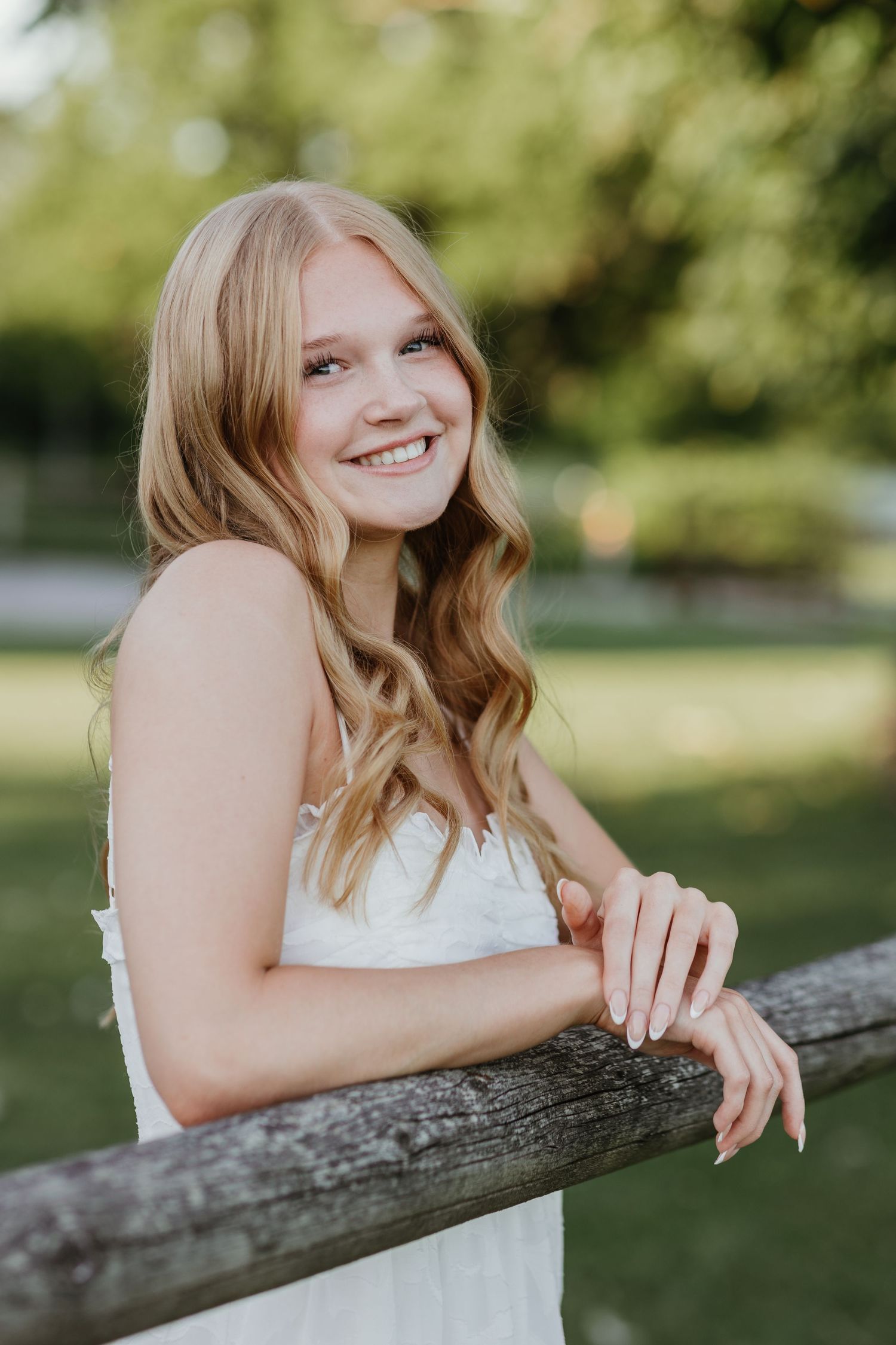 Senior in a white dress leans against a rustic wooden fence on a sunny summer day with blurred green foliage background.