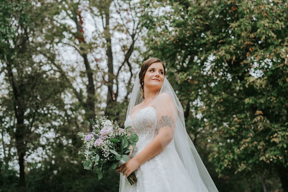 A bride in a white lace gown poses in a garden setting with trees and greenery in the background.