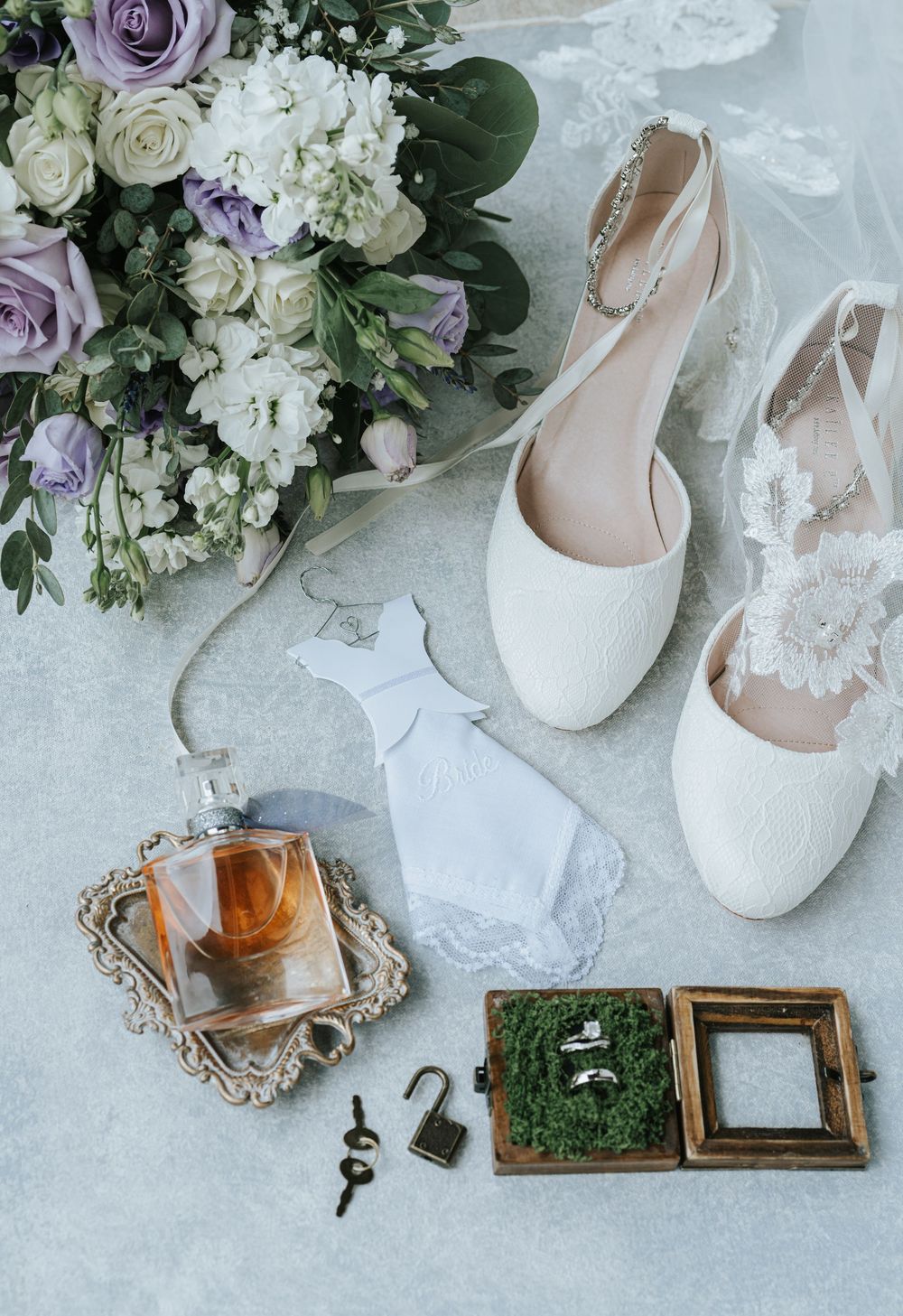 Wedding accessories flatlay with white lace shoes, floral bouquet, antique frames, and decorative items on light surface.