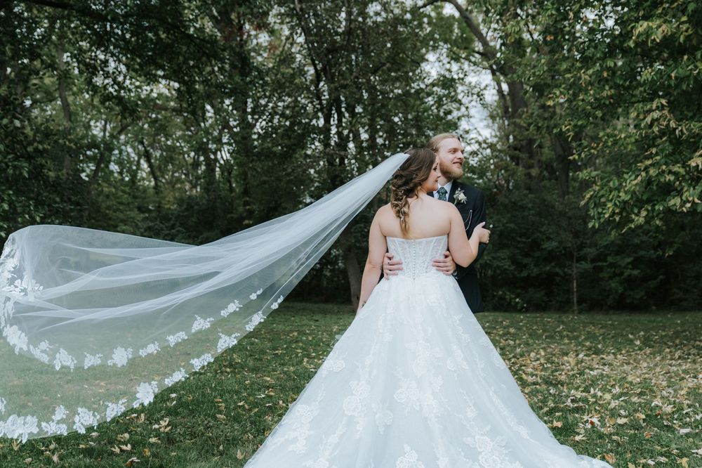 A flowing white wedding veil trails behind a bride in a lush green outdoor garden setting.