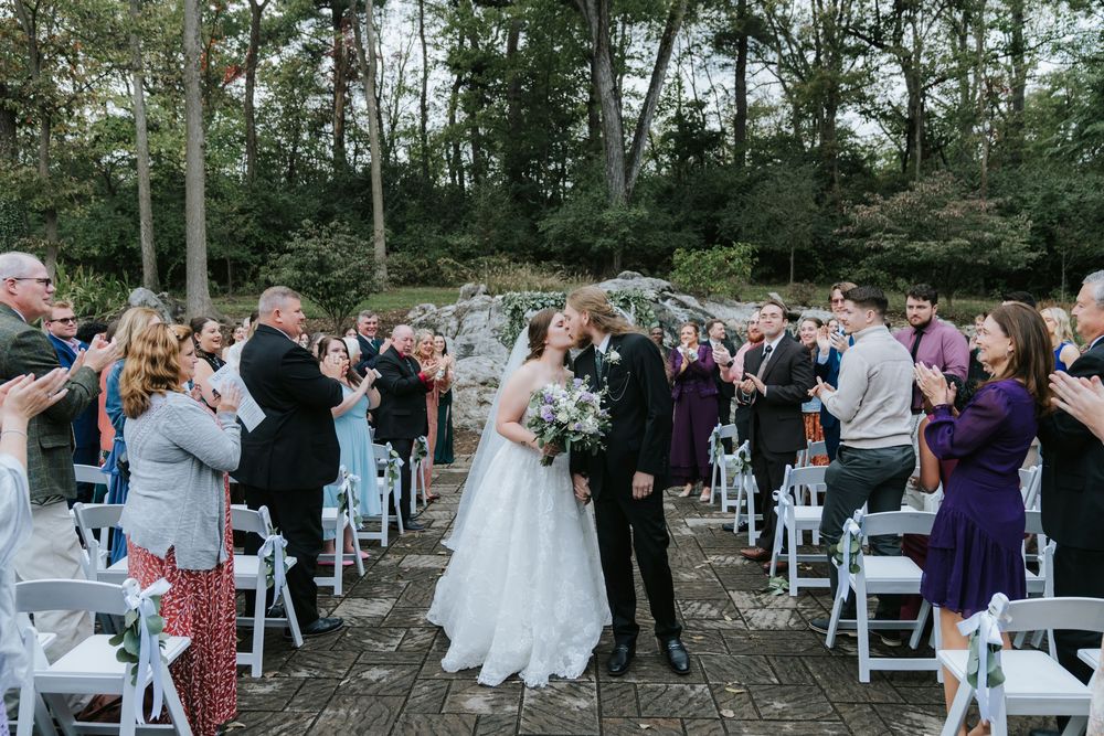 A joyful outdoor wedding ceremony takes place on a stone patio surrounded by trees and white folding chairs.