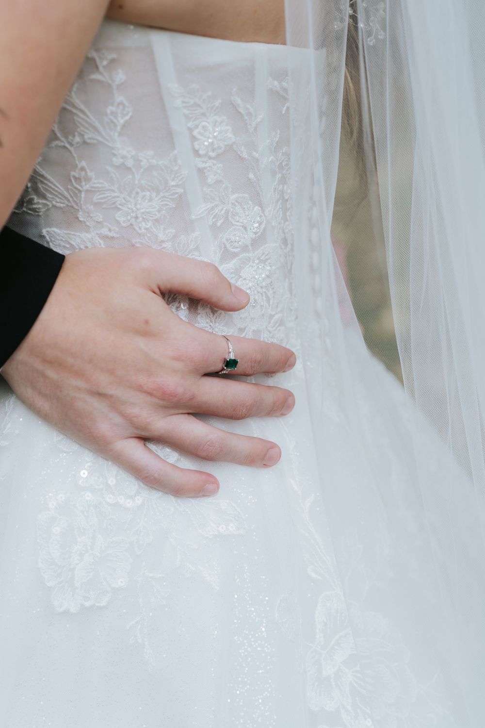 Close-up details of delicate white lace wedding dress fabric with hands showing ring details.