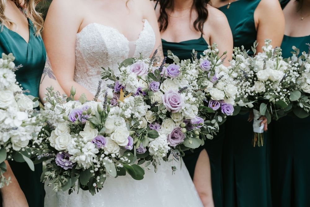 Wedding party holding elegant bouquets with white and purple flowers against emerald green and white dresses.
