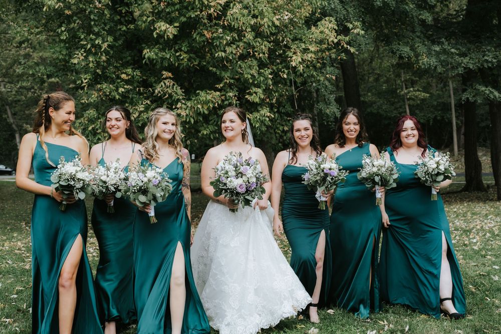 Wedding party in teal bridesmaid dresses poses outdoors in a garden setting with bride in white gown.