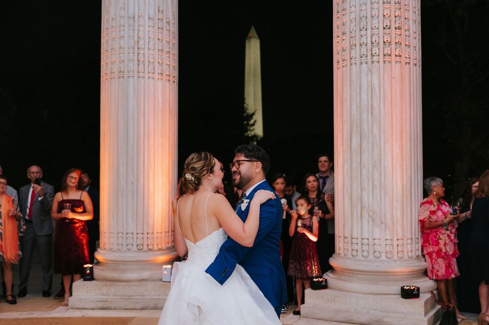 A couple dances between elegant white columns at a formal evening event with guests watching in the background.