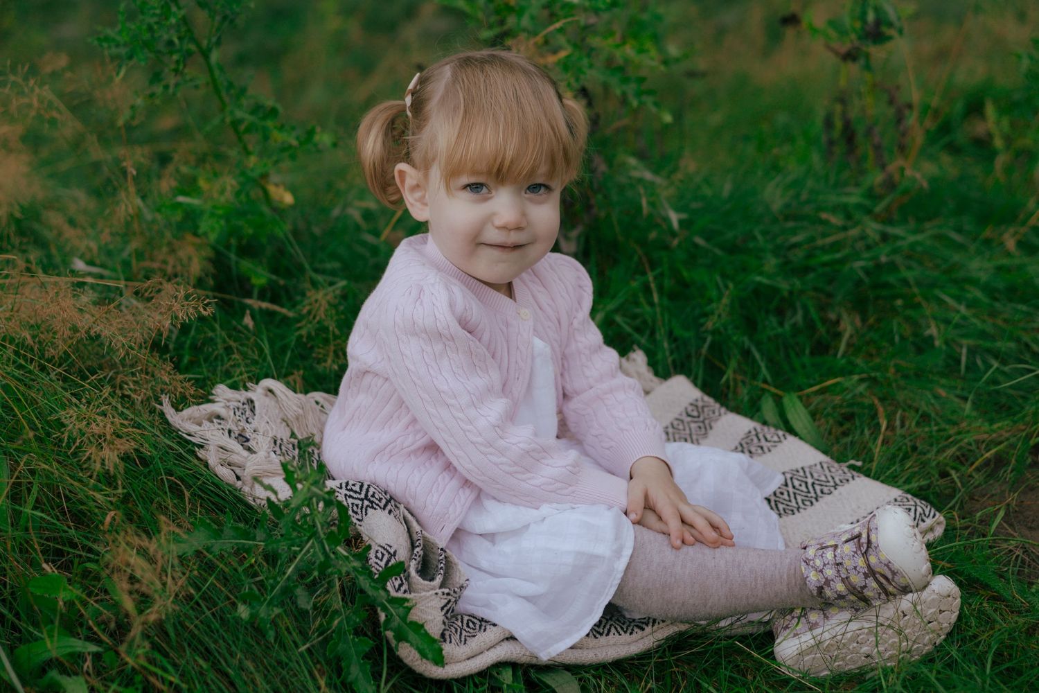 A small child in a pink sweater sitting on a blanket outdoors.