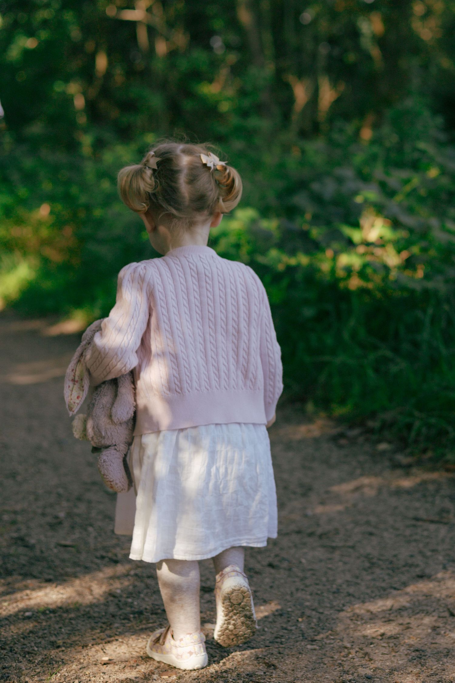 A small child in a pink sweater walking on a forest path while holding a stuffed animal.