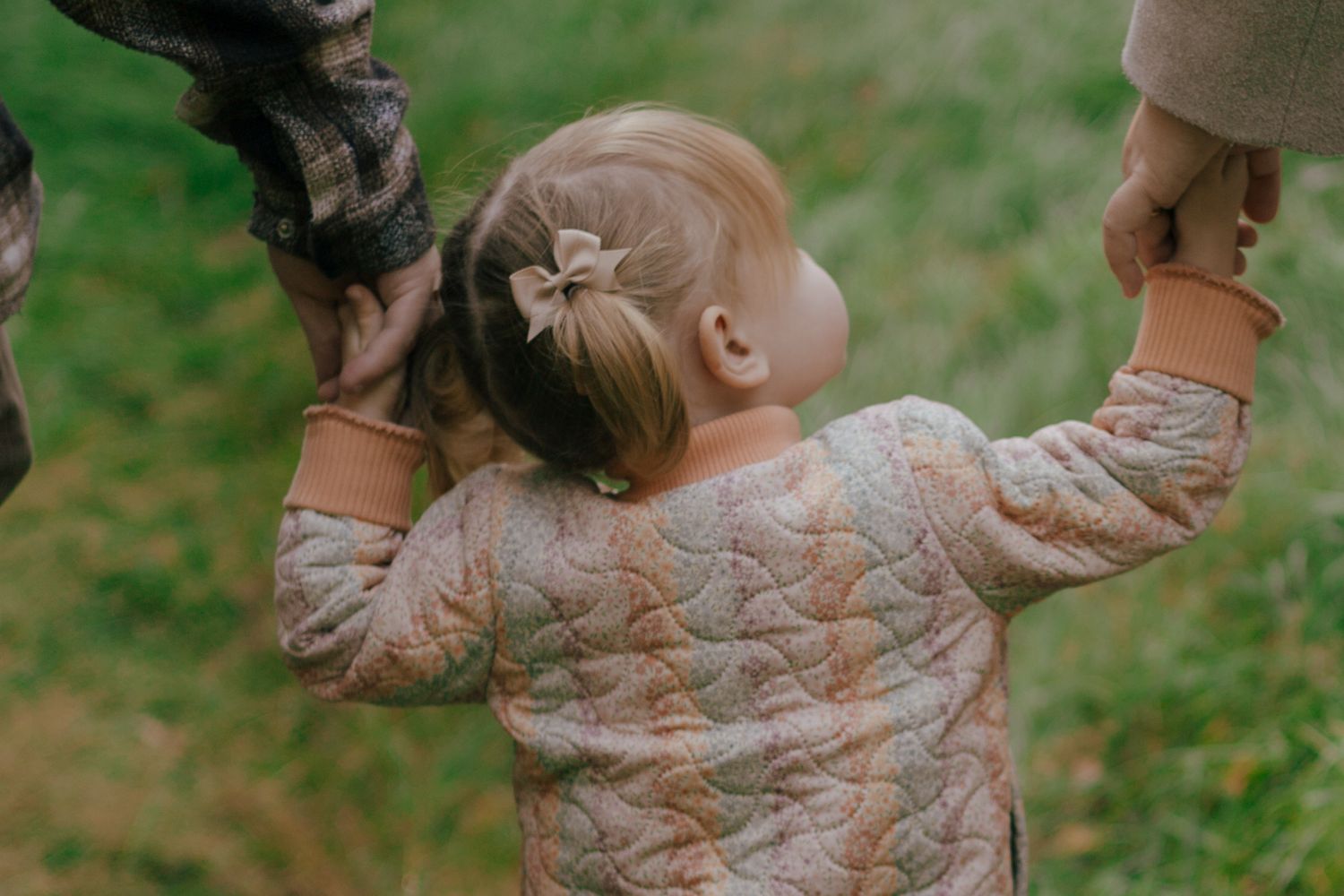 A toddler in a quilted pink jacket reaching up to hold hands with adults.