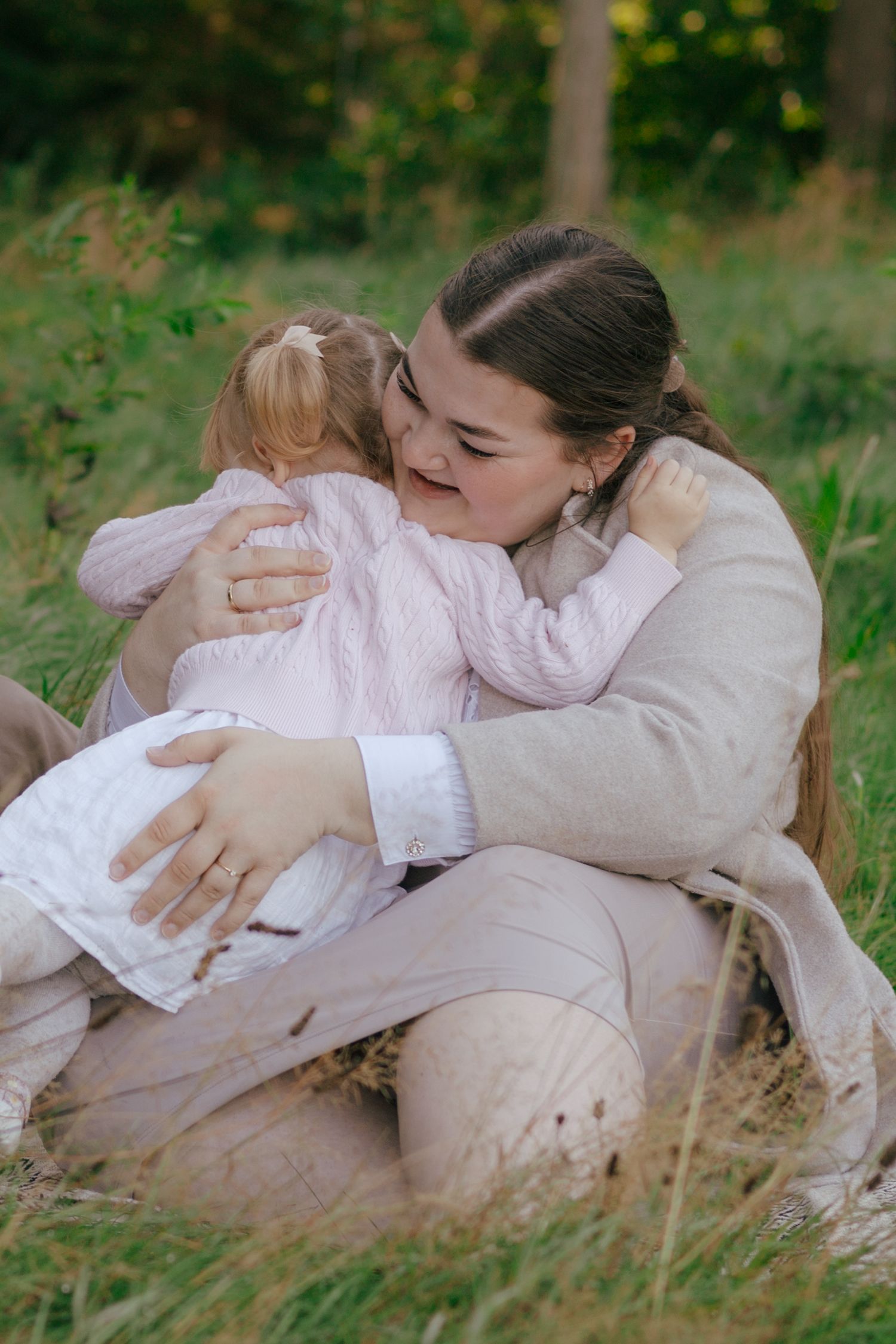 Parent and child share a tender embrace while sitting in tall grass on a sunny day.