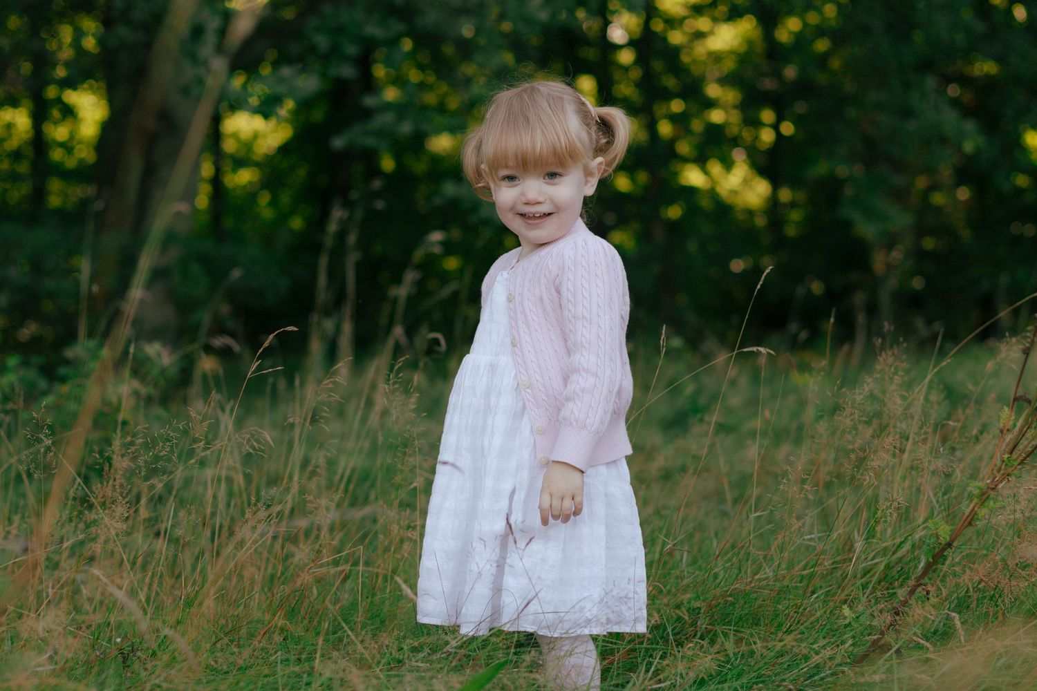 Young child in a white dress standing in a grassy field with trees in the background.