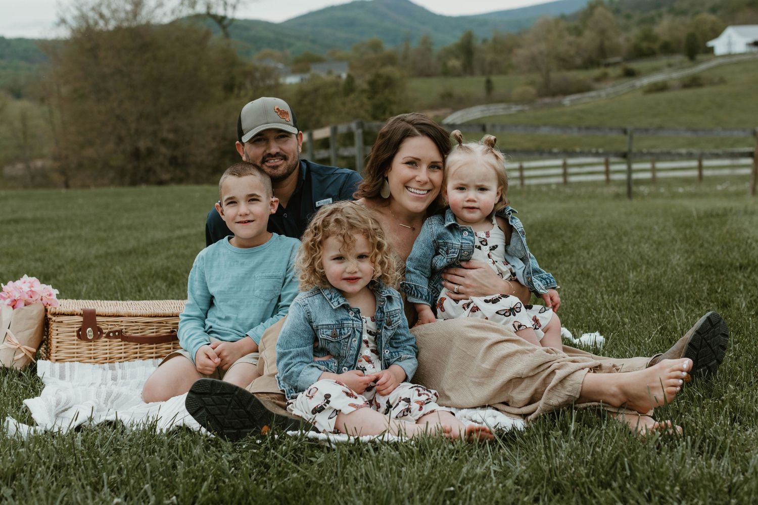 Family having picnic on blanket in grassy field with mountains in background.
