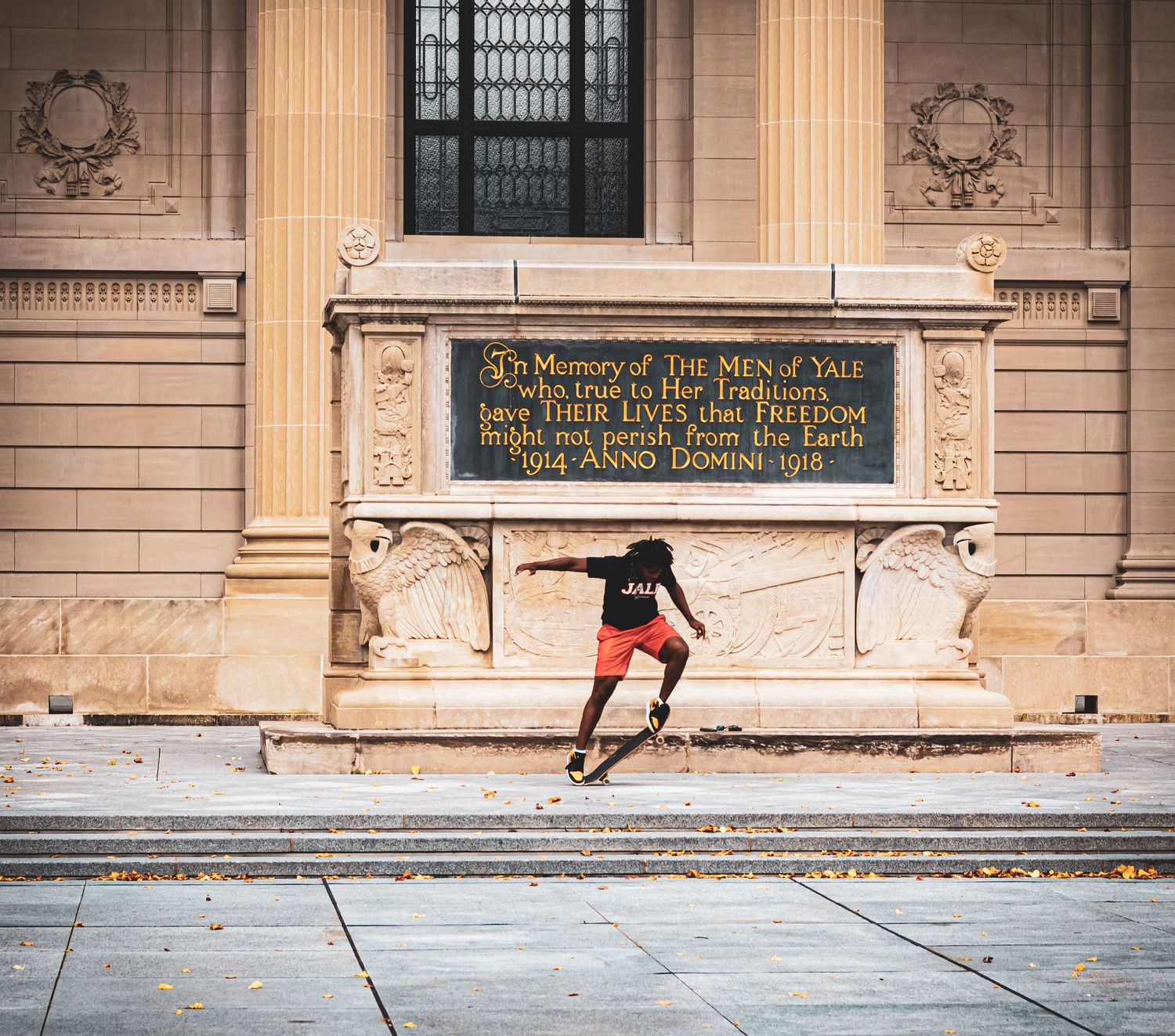 A person in graduation attire doing a skateboard trick in front of an ornate building facade.