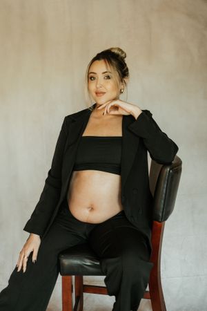 Professional maternity photo of woman in black blazer and crop top sitting on chair.
