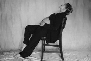 Artistic black and white portrait of person reclining in chair against plain backdrop.