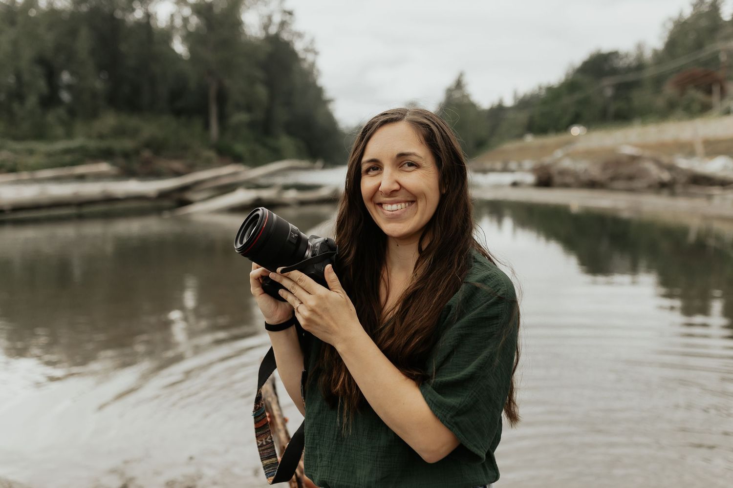 Woman in green shirt holding camera by lake with rocks and trees in background.