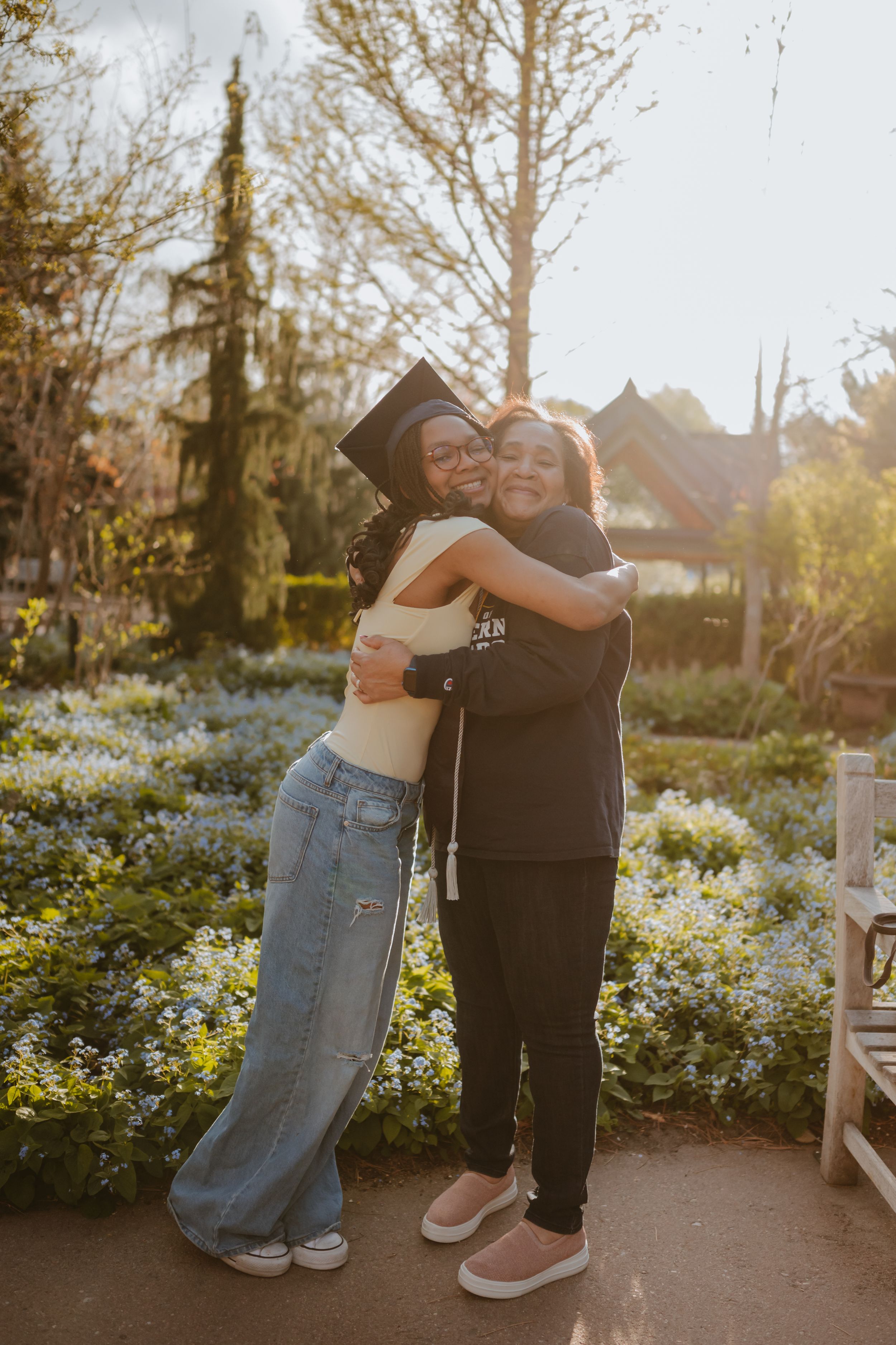 Two people share a warm embrace in a garden at sunset with blooming flowers and trees in the background.
