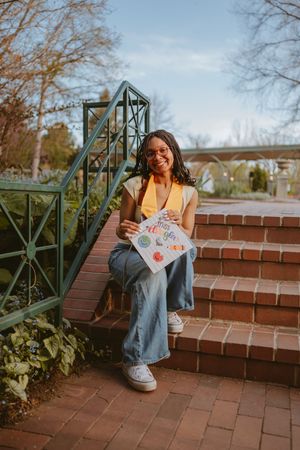 A person in a yellow sweater and jeans sits on brick stairs near a metal railing on an autumn day.