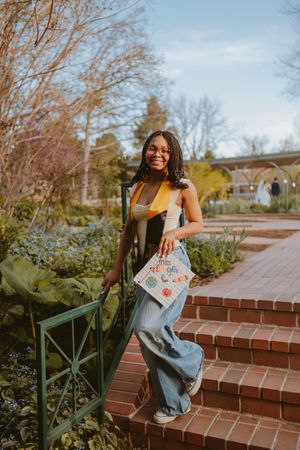A smiling figure in casual attire poses on brick steps in a garden setting during golden hour.