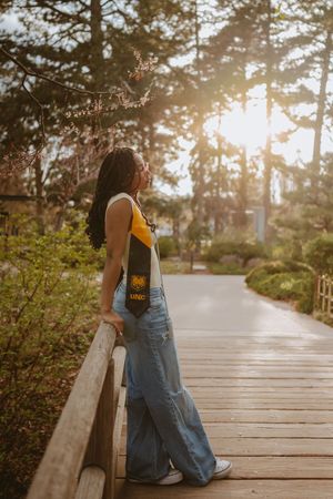 Person in denim overalls sitting alone on a wooden boardwalk during golden hour sunset among trees and foliage.