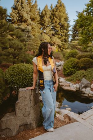 Someone in overalls sits on stone blocks beside a scenic garden pond with evergreen trees.
