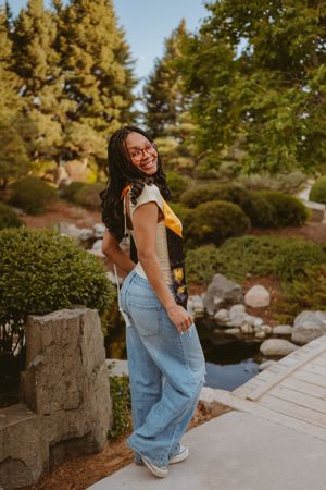 A person in denim overalls poses playfully in a garden setting with evergreen trees and stone features.