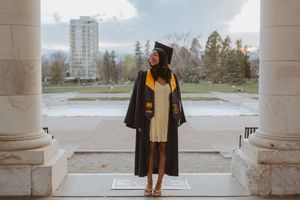 A graduate wearing a black cap and gown poses in front of white stone columns on an overcast day.