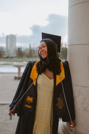 A graduate in yellow and black university attire poses joyfully between white pillars near a waterfront.