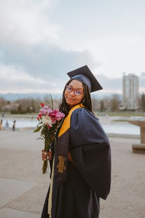 A graduation photoshoot featuring pink flowers and a traditional black cap and gown against an overcast sky.