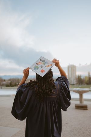 A graduate in dark blue cap and gown holds up their diploma against a cloudy sky by the water.