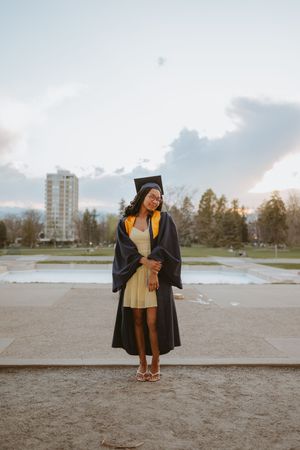 Graduate in academic regalia poses against sunset sky near urban waterfront.