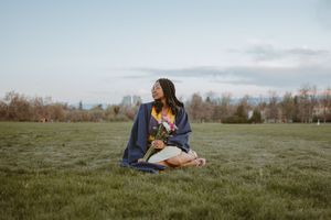 Someone sits cross-legged in a denim jacket on grassy field at sunset with city skyline in background.