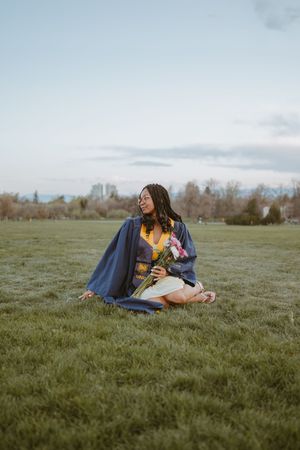 A graduate relaxes on grassy field with diploma and flowers while wearing traditional academic regalia.