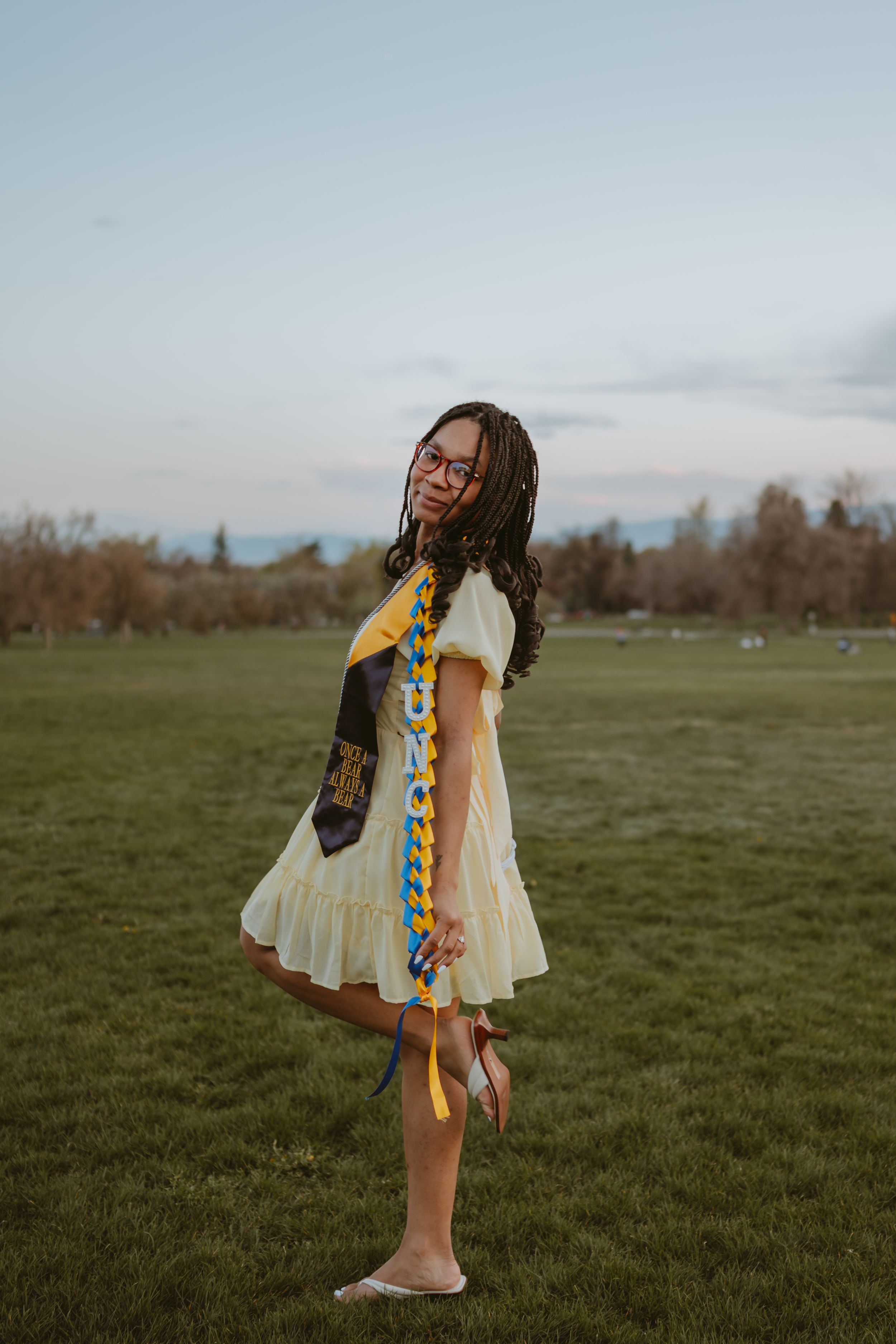 A graduate in a white dress and decorated stole twirls in a grassy field.