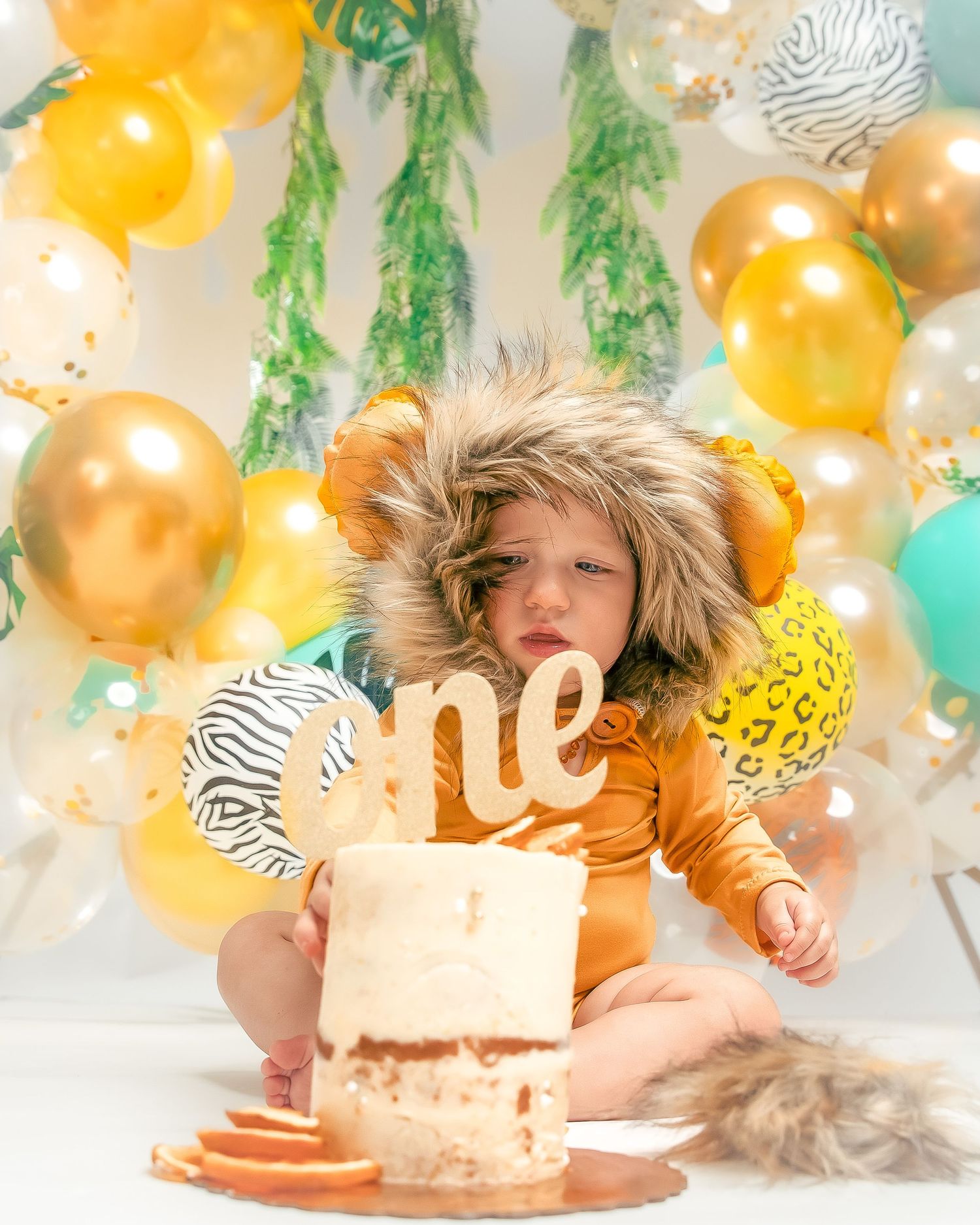 An adorable baby in a lion costume celebrating their first birthday with a smash cake and balloons.
