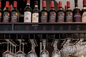 Wine bottles displayed on shelf above hanging stemware glasses in bar setting.