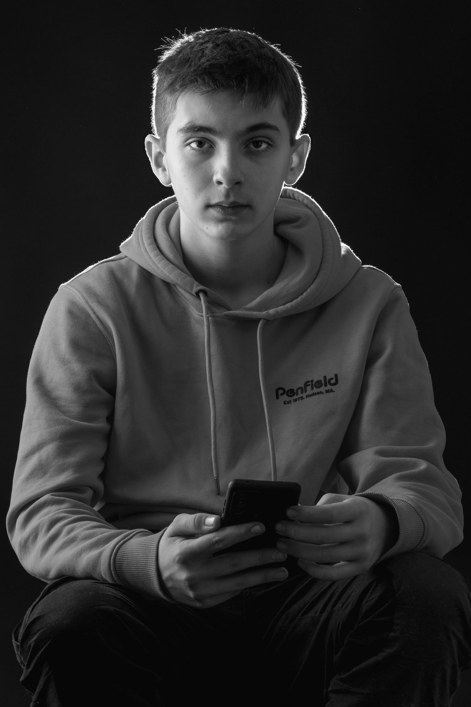 A moody black and white portrait of a person in a hoodie holding a phone.
