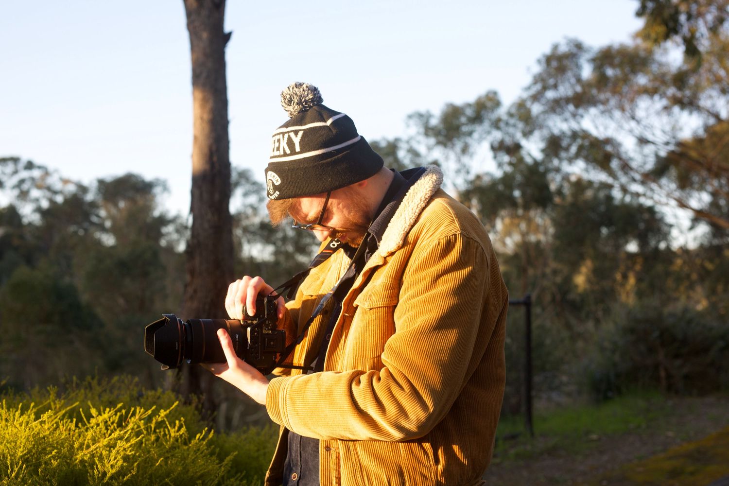 Person in yellow jacket and beanie examines camera during golden hour outdoor photoshoot.