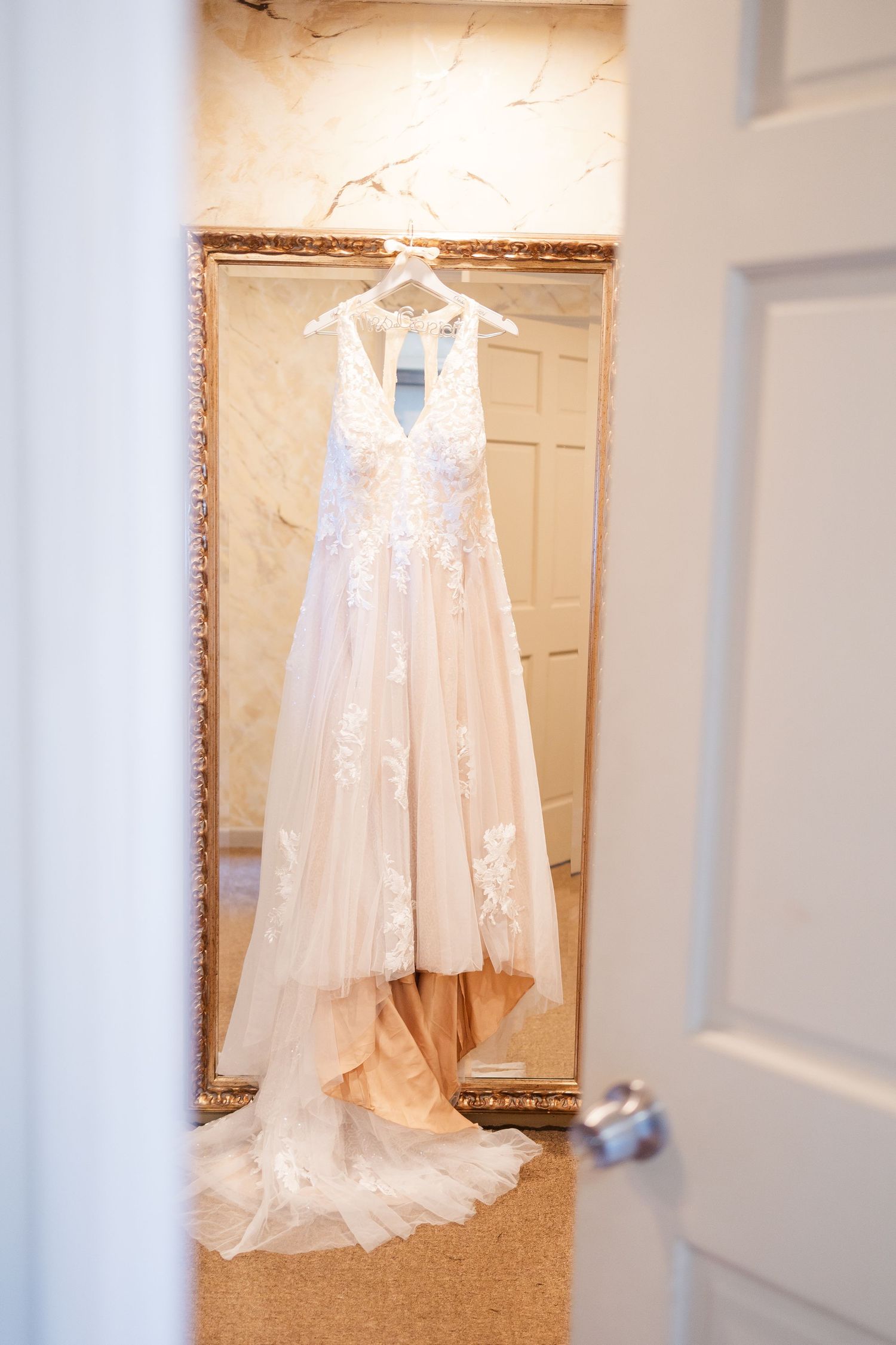White lace wedding dress hanging in doorway against cream colored wall.
