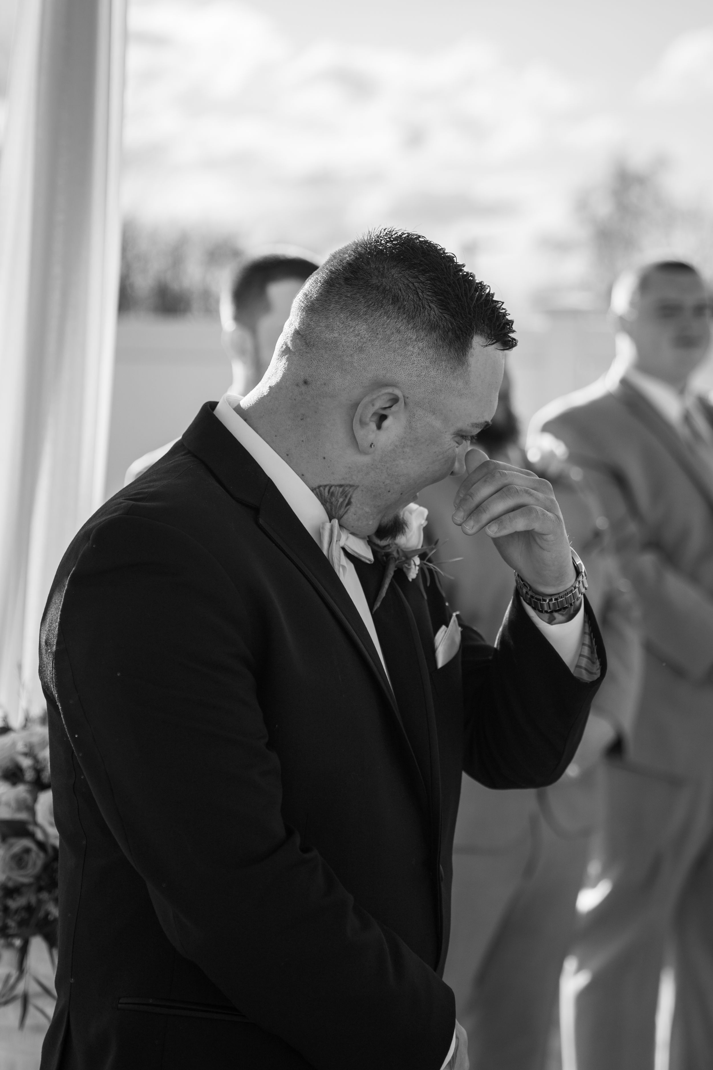 Black and white photo of groom in formal suit showing emotional reaction during wedding ceremony.