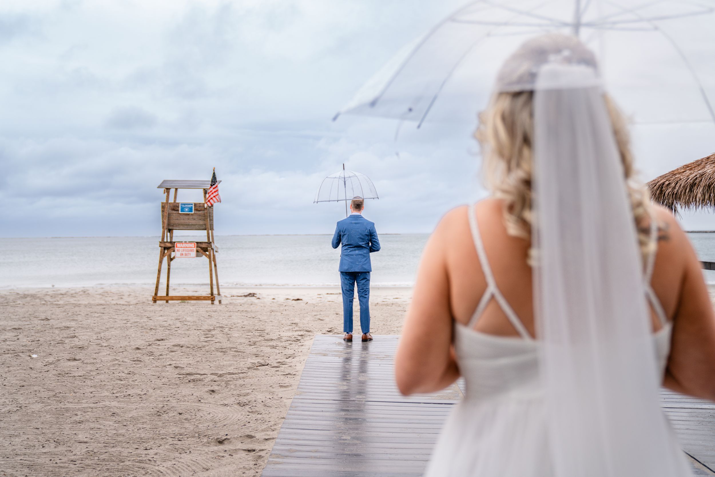 Rainy beach wedding ceremony with lifeguard chair and umbrella on overcast day.