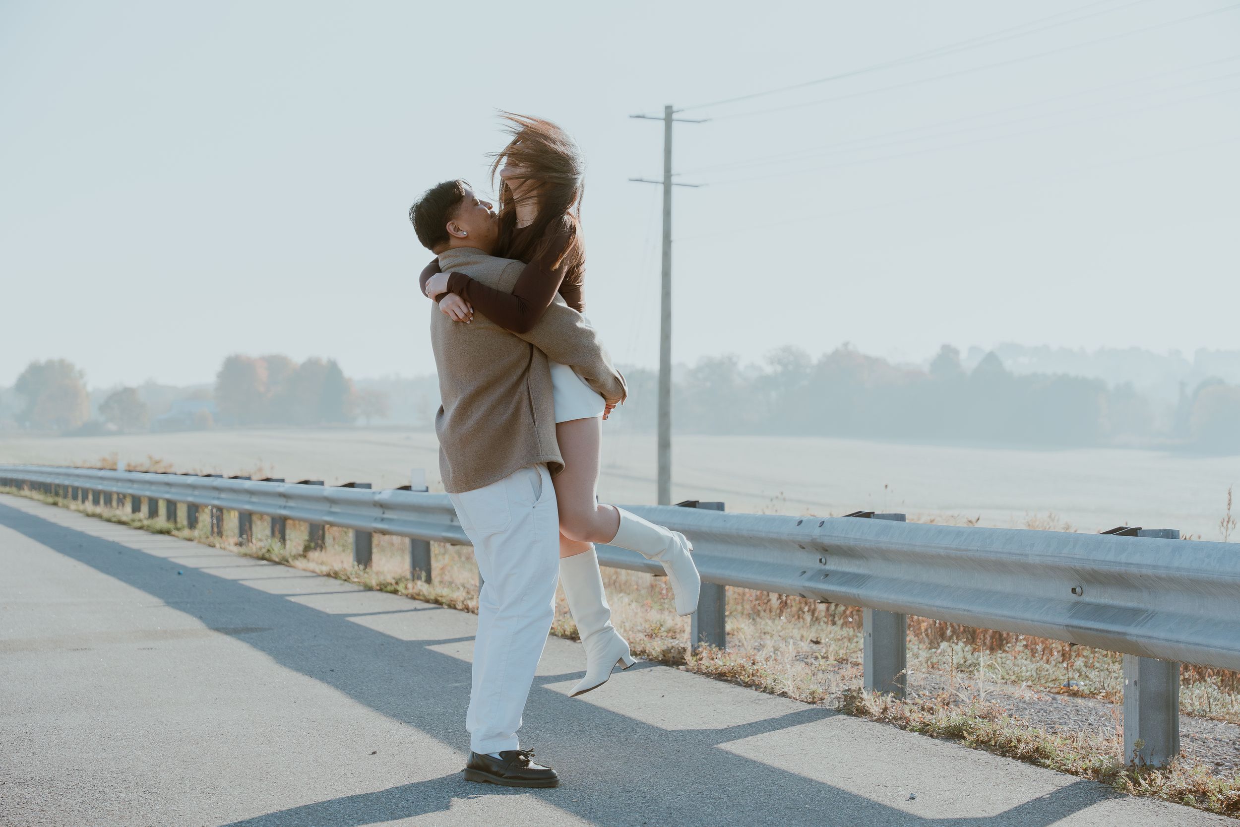 Playful couple embracing on a guardrail beside a rural road.