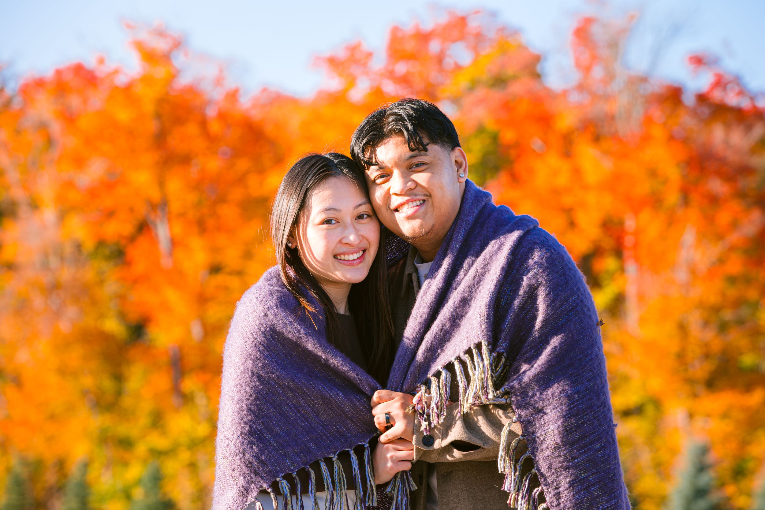 A couple wrapped in matching blankets pose together against vibrant orange autumn foliage in the background.