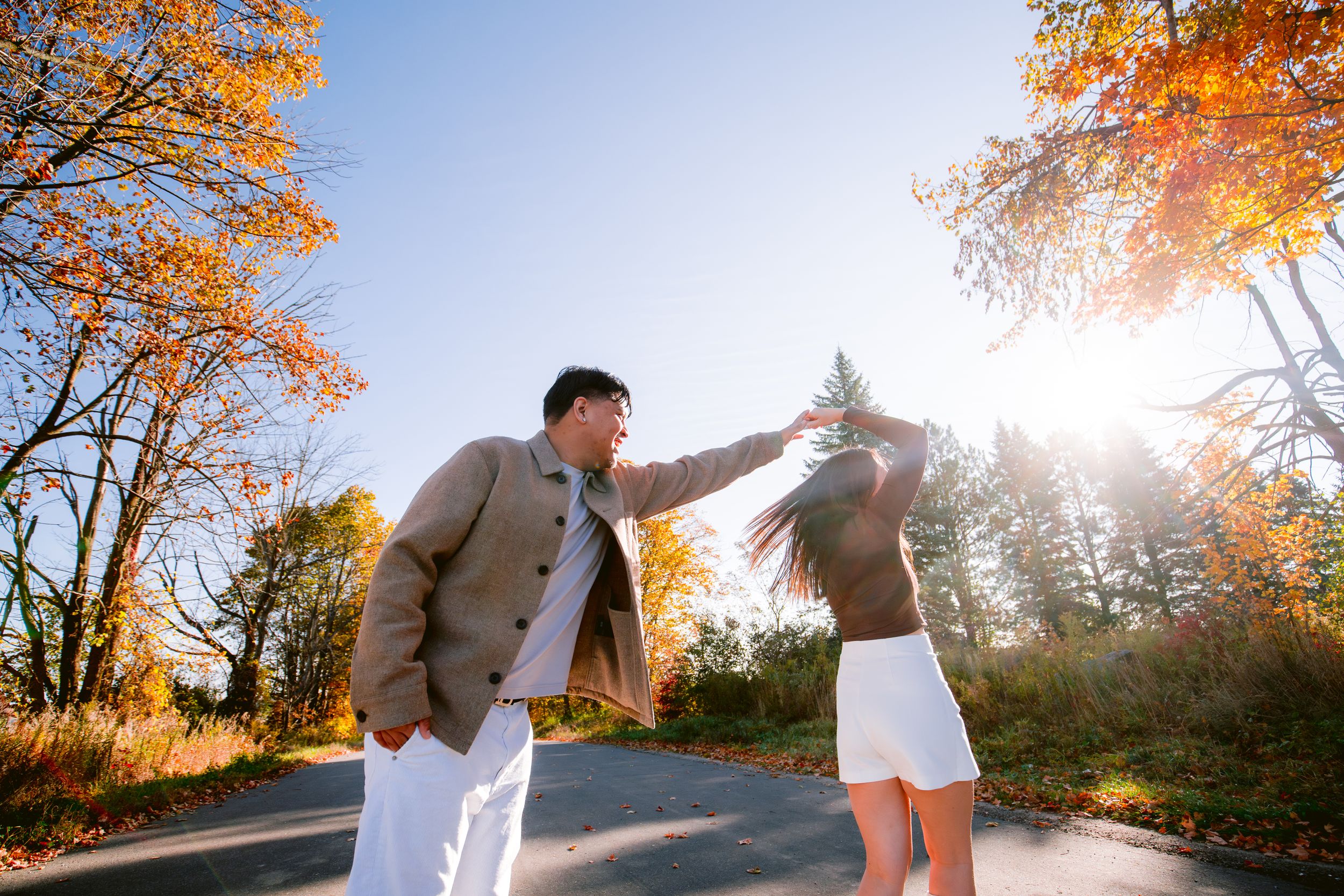 A joyful couple holds hands and dances together on a path surrounded by autumn trees.