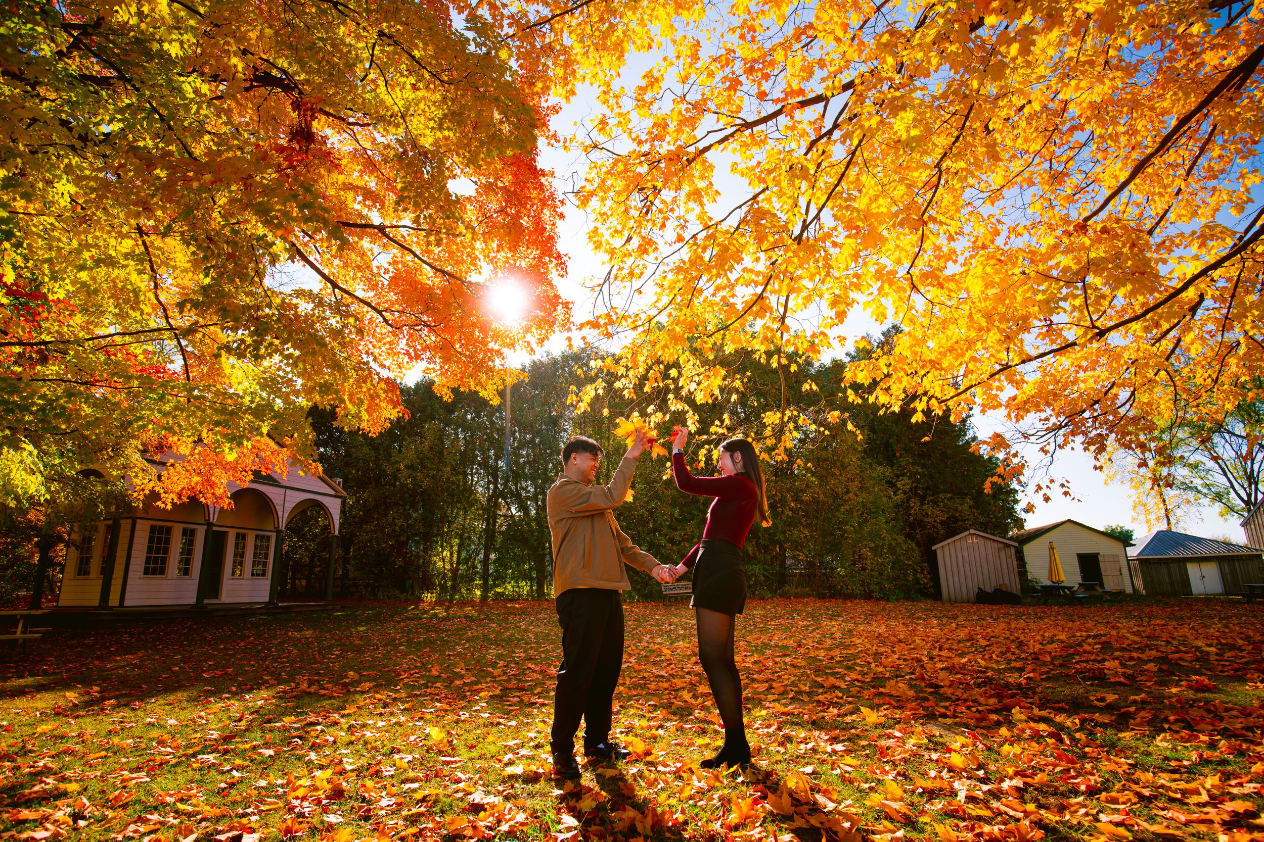 Two people jump joyfully under vibrant orange maple trees on an autumn day.