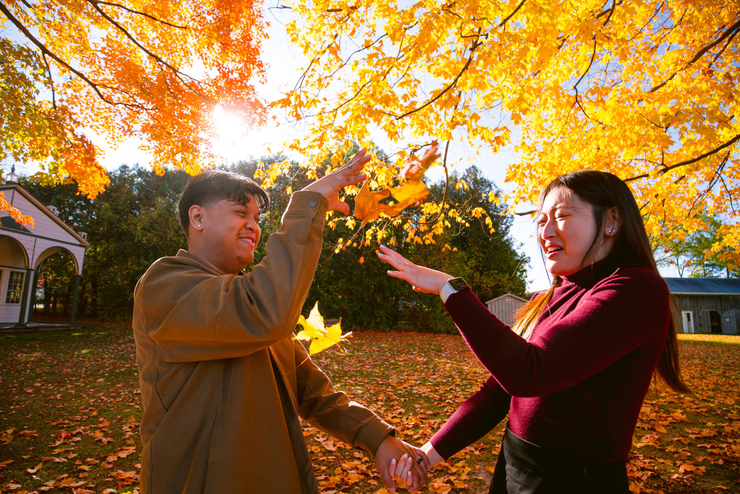 A couple plays with falling autumn leaves under bright orange maple trees.