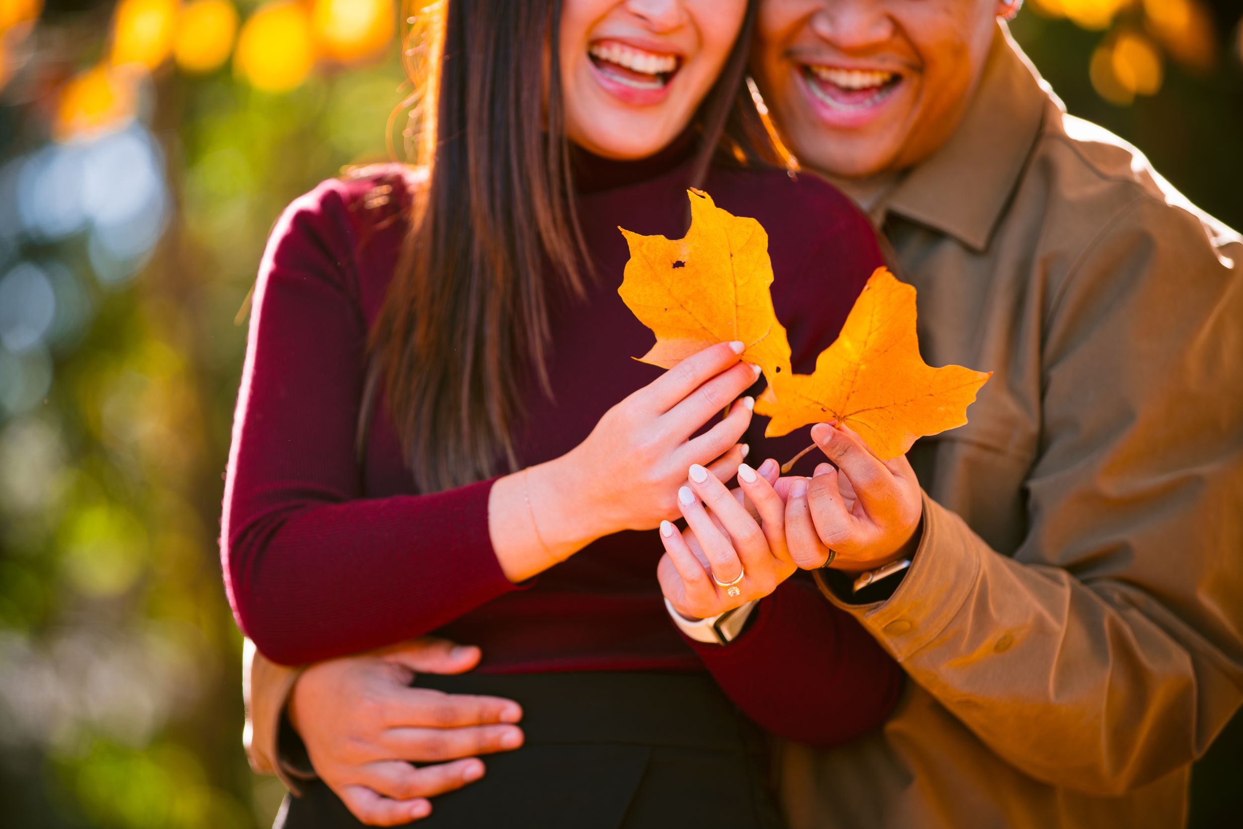A couple holding bright yellow autumn maple leaves against a backdrop of golden bokeh lights.