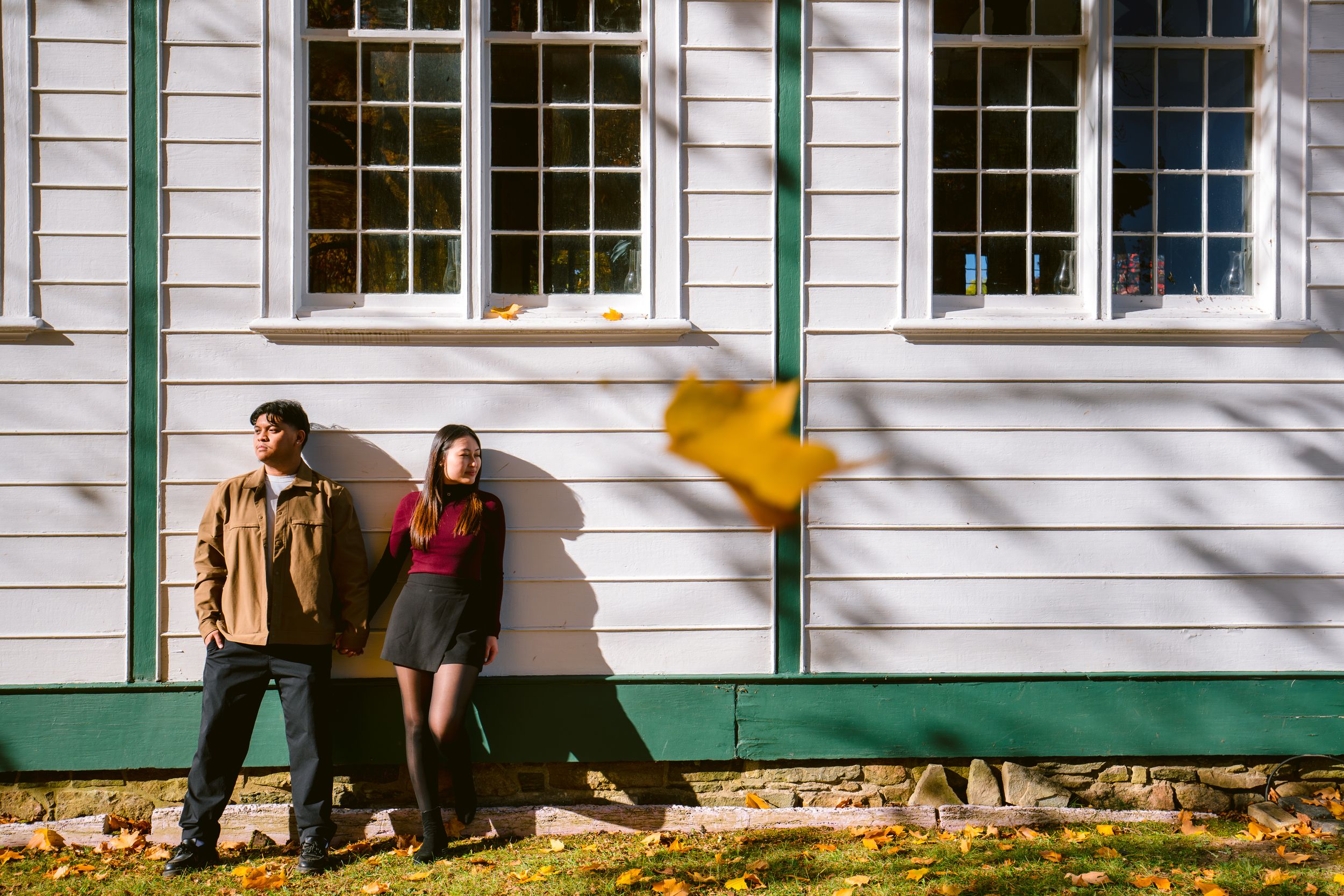 A couple standing against a white building with green trim as yellow leaves fall around them.