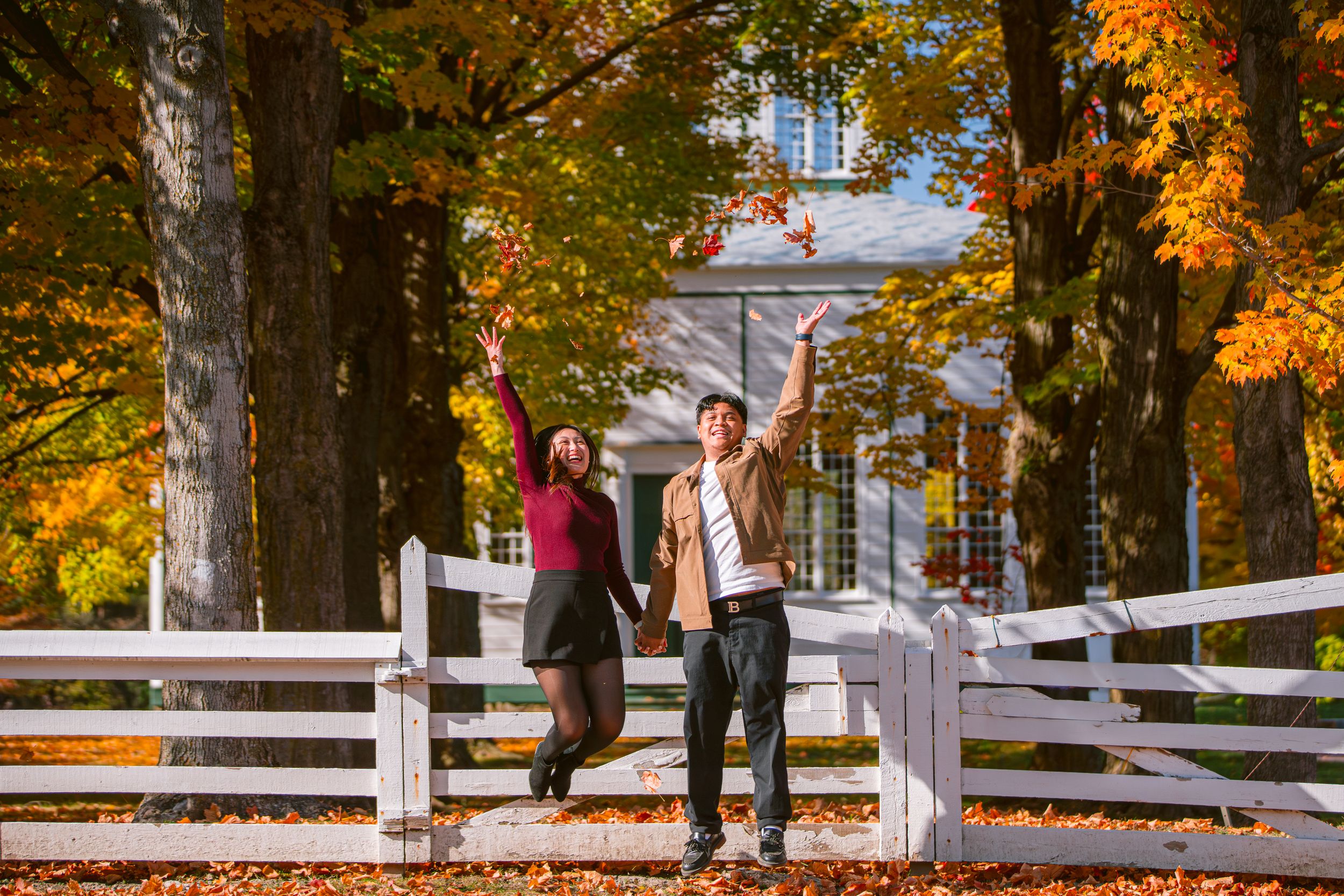 People enjoy a playful moment on a white fence surrounded by fall foliage at a historic property.
